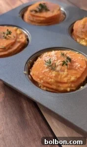 Close-up of a muffin tin filled with Sweet Potato Stacks—layers of sliced sweet potatoes topped with herbs and grated cheese—ready for baking on a wooden surface.