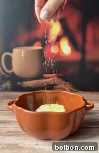 A hand sprinkles salt over a brown dish of Sweet Potato Stacks garnished with a slice of lemon, while a blurred background reveals a mug and the warm glow of a fireplace.