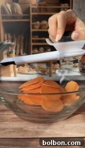 A hand uses a white mandoline slicer to cut thin orange sweet potato slices for Sweet Potato Stacks into a clear glass bowl, with a bakery display blurred in the background.