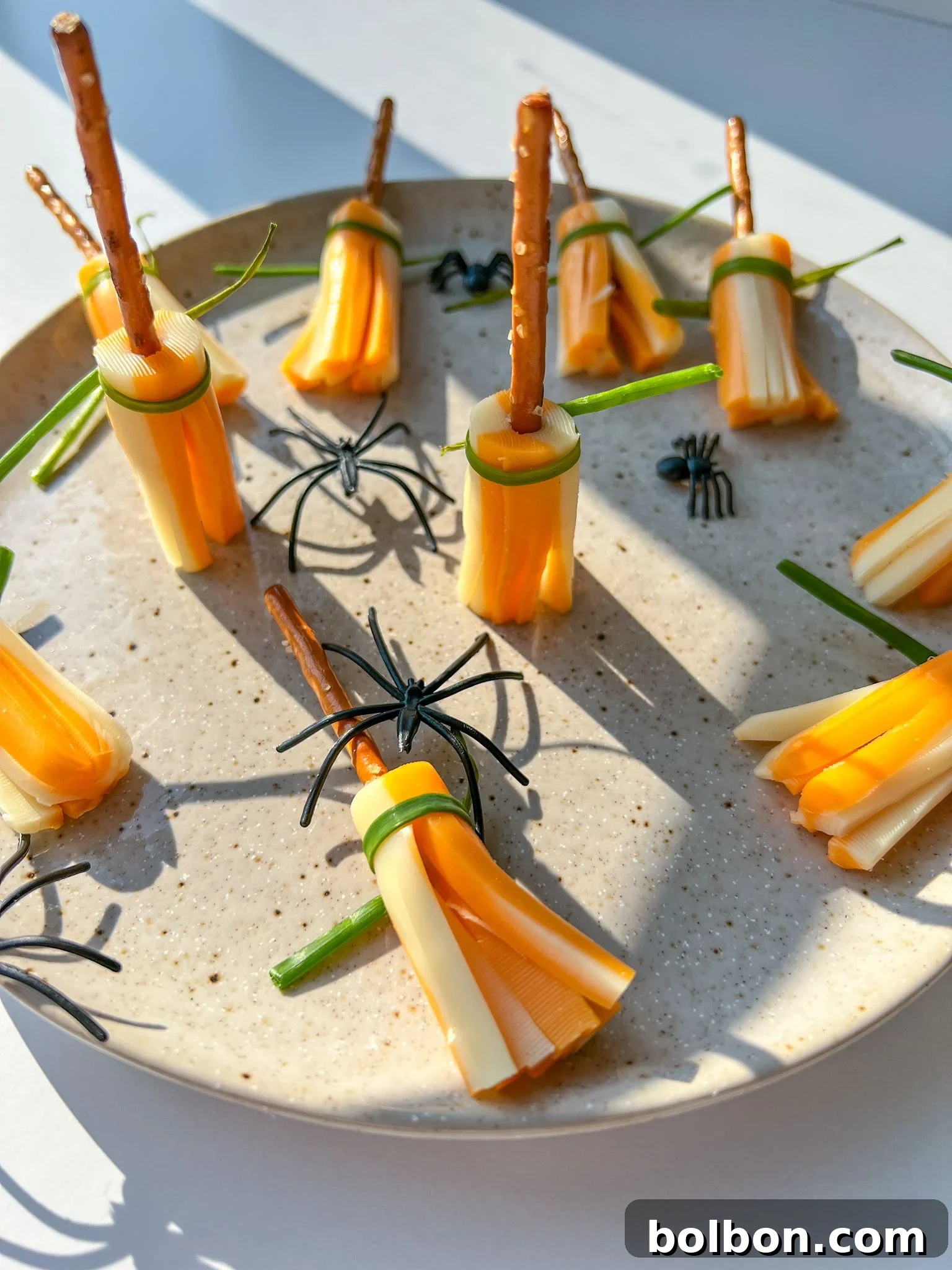 A festive Halloween snack display featuring Cheese Broomsticks made from string cheese and pretzel sticks, adorned with small plastic spiders on a white plate with subtle decor.