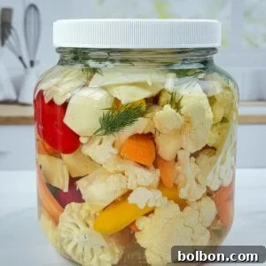 A large glass jar filled with quick pickled veggies—cauliflower, carrots, and peppers—sits on a white countertop in a bright kitchen setting.