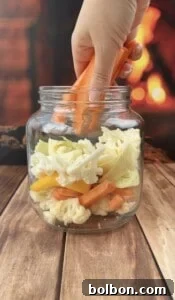 A hand places carrot sticks into a glass jar of quick pickled veggies, layered with cauliflower, yellow pepper, and cabbage. The background features a warm, blurred fireplace scene.