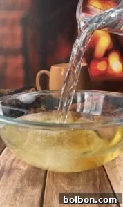 Hot water is being poured from a glass pitcher into a clear bowl with lemon slices, perfect for preparing quick pickled veggies. A tan mug and a warm fireplace are blurred in the background, creating a cozy atmosphere.