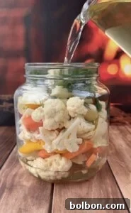 A glass jar filled with quick pickled veggies like cauliflower, carrots, and yellow peppers sits on a wooden surface as clear liquid is poured in, set against a blurred warm-toned background.
