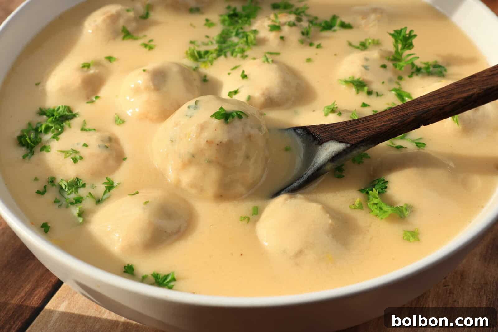 Zesty Lemon Meatballs 2 A bowl of meatballs covered in creamy sauce, garnished with chopped parsley. A wooden spoon rests in the bowl, surrounded by the sauce and meatballs.