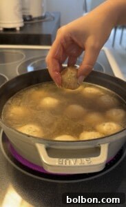 A hand holds a raw meatball above a pot of simmering broth with several meatballs cooking inside on a stovetop.