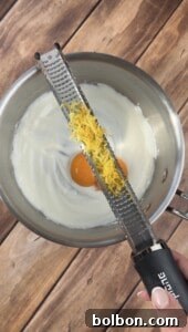A hand holds a zester with fresh lemon zest over a metal bowl containing two egg yolks and white creamy mixture, on a wooden surface.