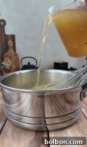 A hand pours golden broth from a measuring cup into a metal saucepan with a whisk inside, set on a wooden surface with kitchen items in the background.