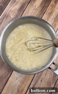 A metal saucepan filled with a light yellow, frothy liquid is being whisked with a metal whisk on a wooden surface.