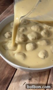Yellow sauce is being poured from a blender into a white bowl filled with round dumplings or meatballs, all sitting on a wooden surface.