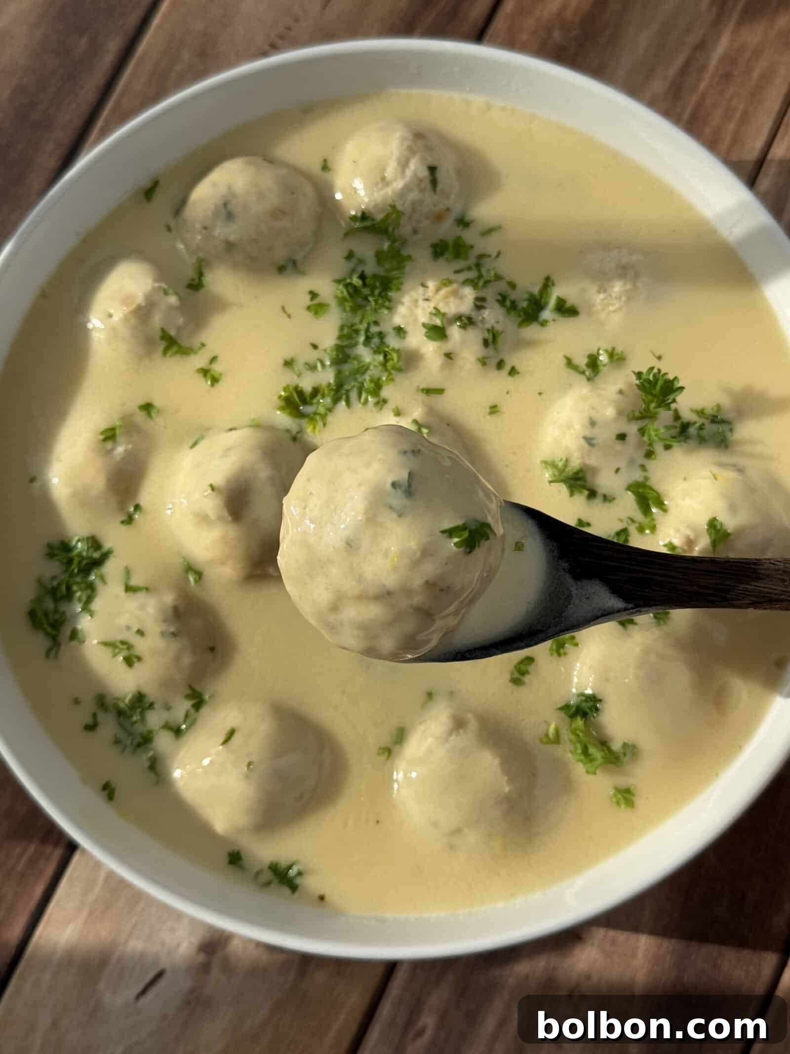 Zesty Lemon Meatballs 3 A bowl of creamy soup with several meatballs, garnished with chopped parsley. A wooden spoon holds one meatball above the bowl. The dish is placed on a wooden surface.