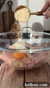 A hand sprinkles breadcrumbs into a glass bowl containing ground meat, an egg, and seasoning, preparing to mix ingredients on a wooden countertop in a kitchen setting.