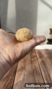 A close-up of a hand holding a round, uncooked dumpling or meatball mixture with herbs, above a wooden surface in natural sunlight.