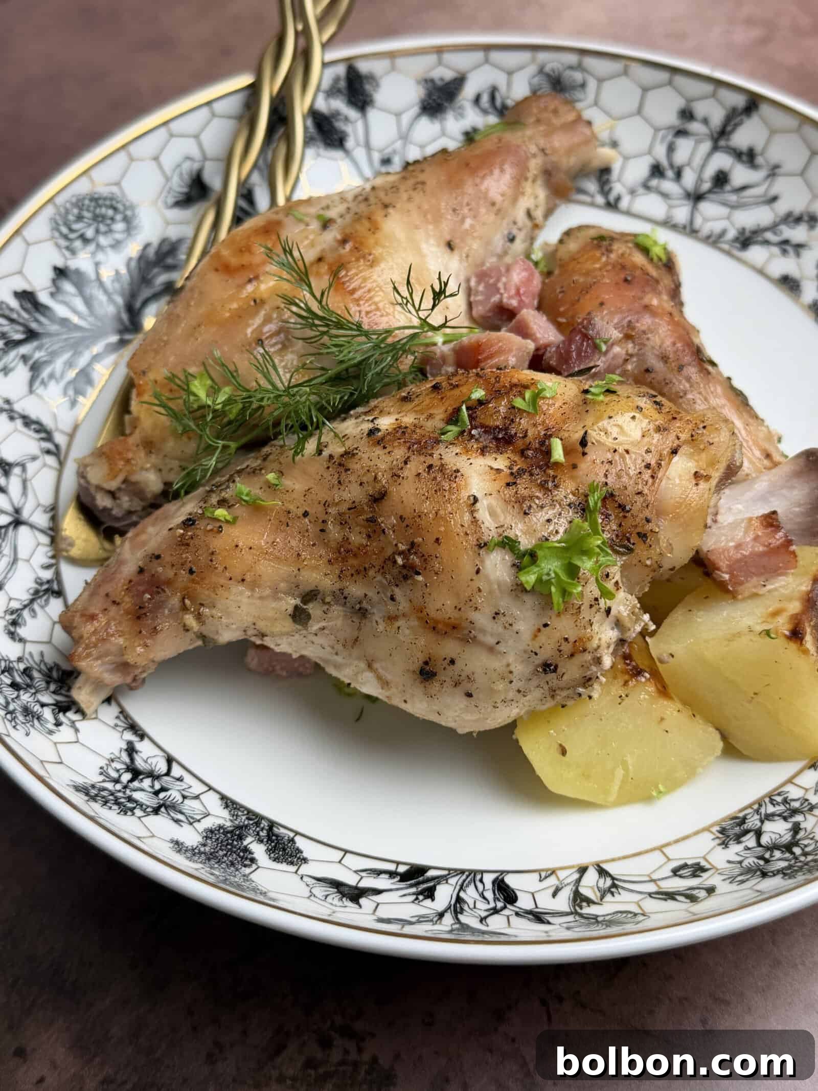 Rustic Baked Rabbit 2 A close-up of a plate with succulent, golden-brown roasted rabbit pieces, garnished with fresh herbs, served alongside crispy roasted potatoes and small cubes of savory pancetta on an elegant, floral-patterned dish. This gourmet meal promises a delightful experience, perfect for those seeking tender baked rabbit recipes.