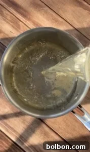 A clear liquid is being poured from a glass container into a stainless steel pot on a wooden surface, with light reflections on the water and pot.