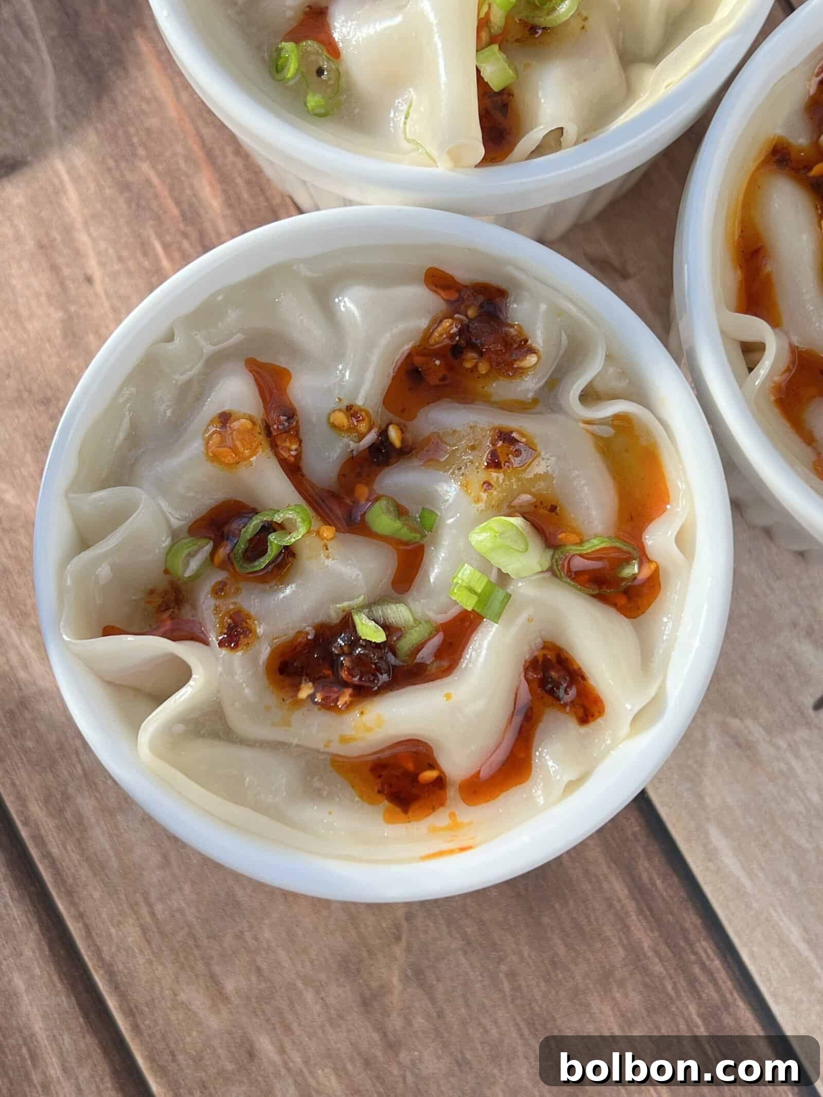 Shrimp Dumpling Lasagna: A white bowl filled with steamy, layered shrimp and dumpling wrappers, generously topped with glistening chili oil and freshly chopped green onions, resting on a rustic wooden surface.