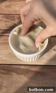 A hand gently presses a thin sheet of dough into a small white ramekin on a wooden surface, carefully shaping and submerging the dough to fit the contours of the dish.
