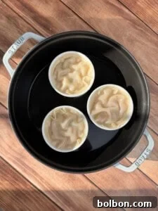 Three white ramekins, each filled with uncooked, wavy dough and layered ingredients, are carefully arranged within a round black Staub pot containing a small amount of water. The pot sits on a wooden surface, with sunlight casting gentle shadows.