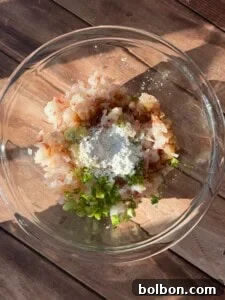 A glass bowl containing precisely chopped shrimp, vibrant green onions, a mound of white cornstarch, and various seasonings, all arranged on a rustic wooden surface under natural sunlight.