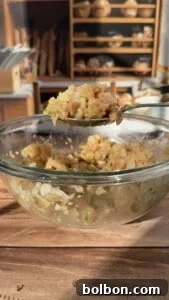 A glass bowl filled with a well-mixed shrimp and vegetable blend, appearing uniform in texture and color, sits on a wooden table. A spoon holds a portion of the mixture, indicating its consistency. Shelves with loaves of bread are visible in the blurred background.