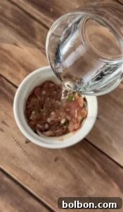 A clear glass is pouring water into a white bowl filled with chopped raw meat and herbs, placed on a wooden surface.