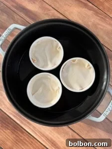 Three white ramekins covered with dough sit in a black round baking dish filled with water, placed on a wooden surface.