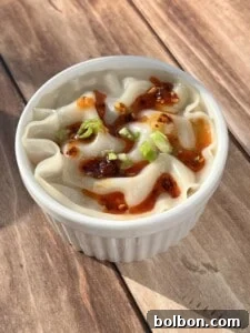 A white ramekin filled with steamed dumplings, garnished with chopped green onions and drizzled with chili oil, sits on a wooden surface.