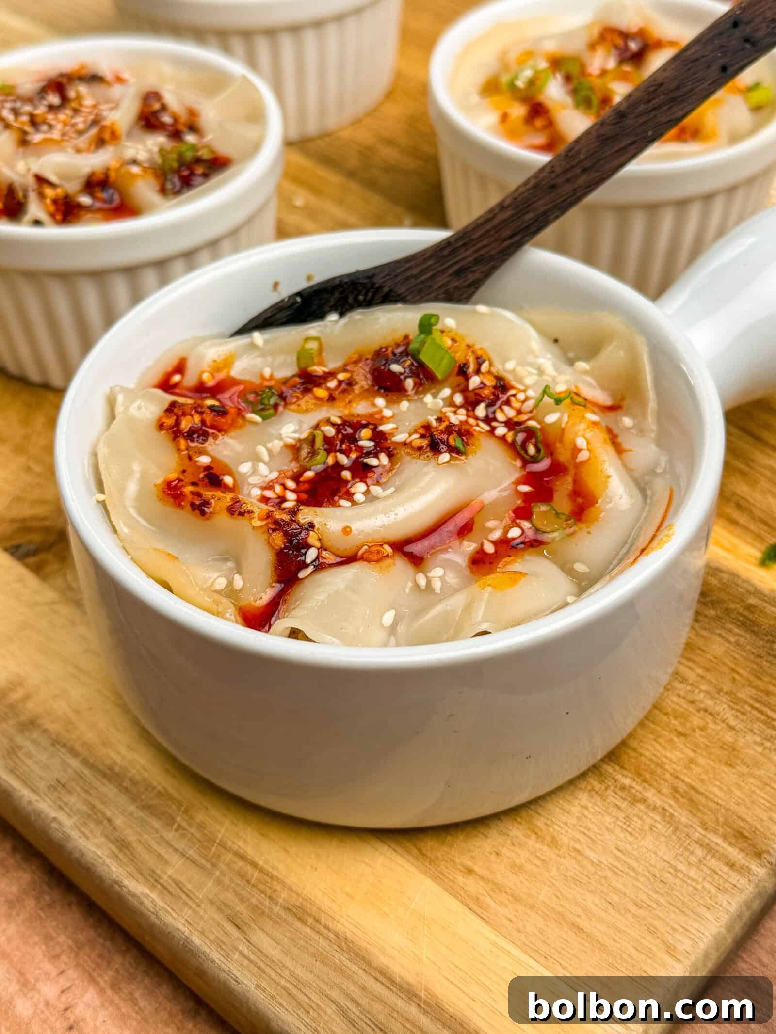 A white ramekin filled with chili oil dumplings, inspired by the VIRAL PORK DUMPLING LASAGNA trend, is garnished with sesame seeds and green onions on a wooden board. A dark spoon rests on the edge, with more dumplings in the background.