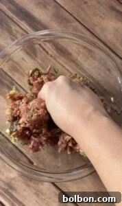 A hand mixes ground meat with chopped green herbs and seasonings in a clear glass bowl on a wooden surface.