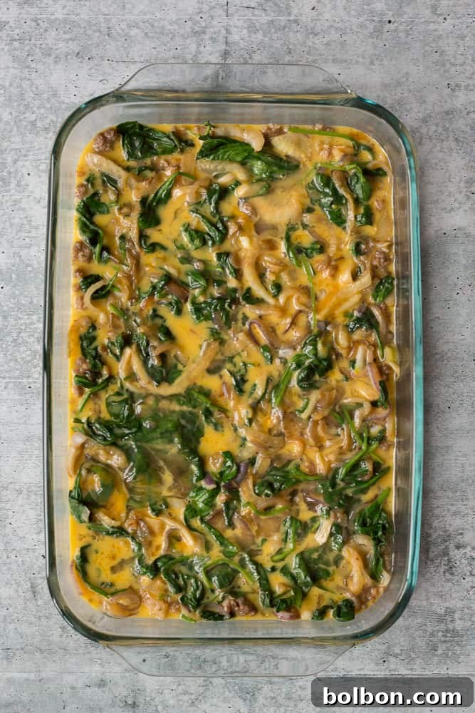 Whisked egg mixture being poured evenly over layers of sausage and vegetables in a 9x13 baking dish, completing the assembly of the make-ahead casserole.
