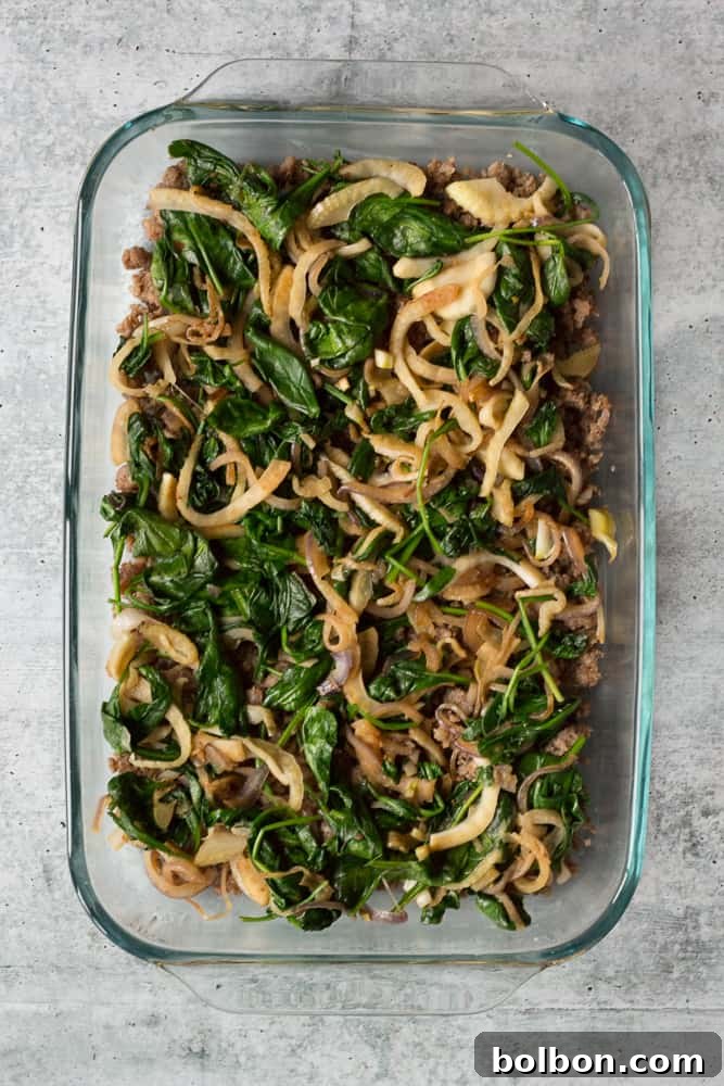 Sautéed onion, fennel, and wilted spinach mixture spread evenly over the sausage layer in a 9x13 baking dish for the casserole.