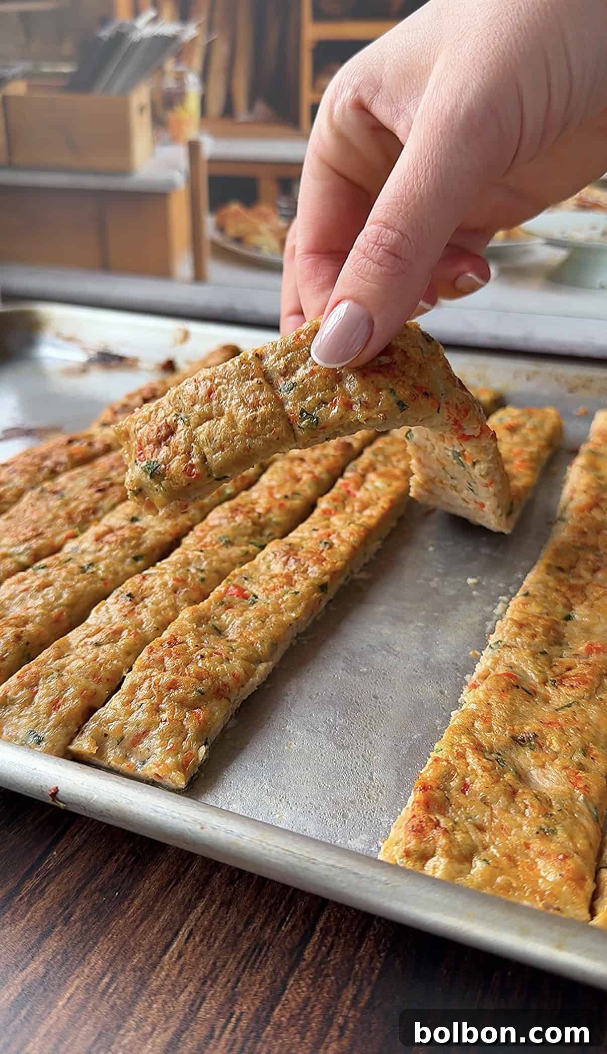 A hand lifts a rectangular slice of seasoned, baked kebab on a metal baking sheet. 