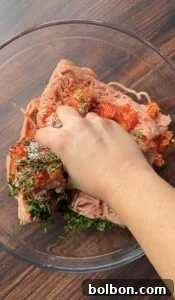 A hand mixing ground meat with chopped herbs, spices, and red seasoning in a glass bowl on a wooden surface.