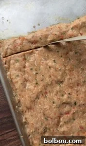 A close-up of a baking tray with a thick, speckled dough mixture being cut into squares with a knife. The dough is flecked with green and red herbs and vegetables.
