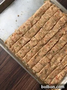 A metal baking tray with evenly cut, uncooked meat mixture bars seasoned with herbs and red pepper, placed on a wooden surface.