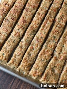 A close-up of a baking tray filled with evenly cut rectangular strips of baked meat or vegetable mixture, showing a golden-brown top with visible herbs and small vegetable pieces.