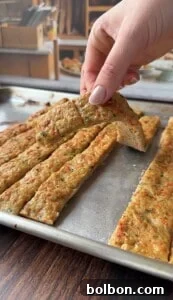 A hand lifts a rectangular slice of seasoned, baked breadsticks from a tray of similar strips on a metal baking sheet. The breadsticks appear golden and speckled with herbs.