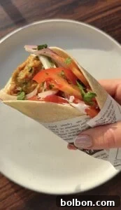 A hand holding a wrap filled with falafel, sliced tomatoes, onions, and fresh herbs, partially wrapped in printed paper, over a white plate on a wooden table.