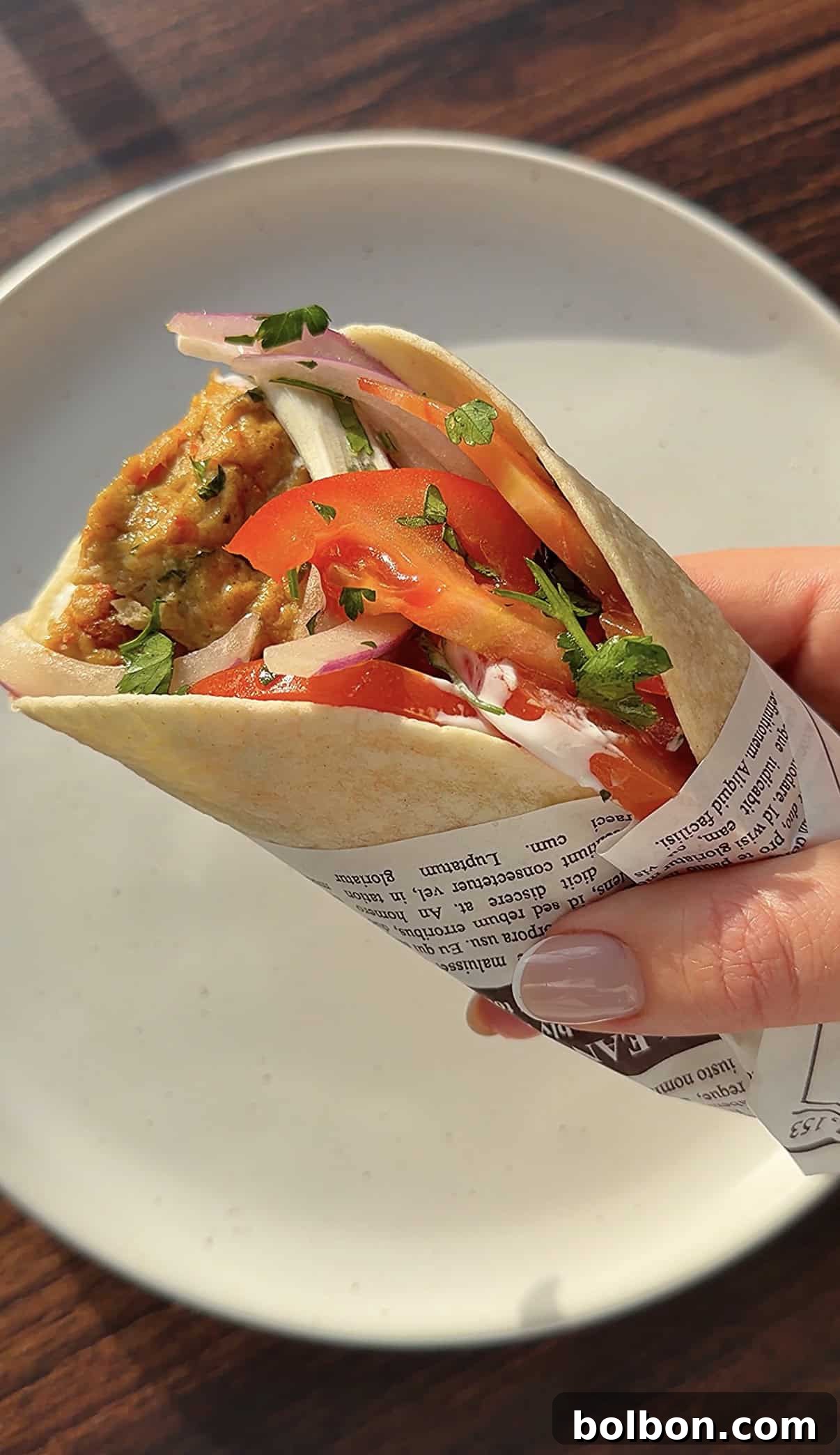 A hand holding sheet pan kebab wrap with sliced tomatoes, onions, and fresh herbs, partially wrapped in printed paper, over a white plate on a wooden table.
