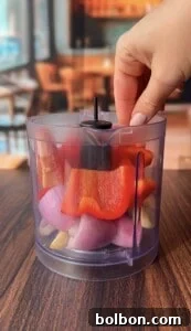 A hand holds the lid of a clear plastic food chopper filled with chopped red bell pepper, red onion, and garlic cloves on a wooden table, with a blurred indoor background.