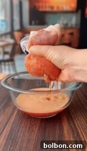 A hand squeezes a mesh bag filled with blended tomatoes over a glass bowl, extracting tomato juice onto a wooden table in a cozy kitchen setting.