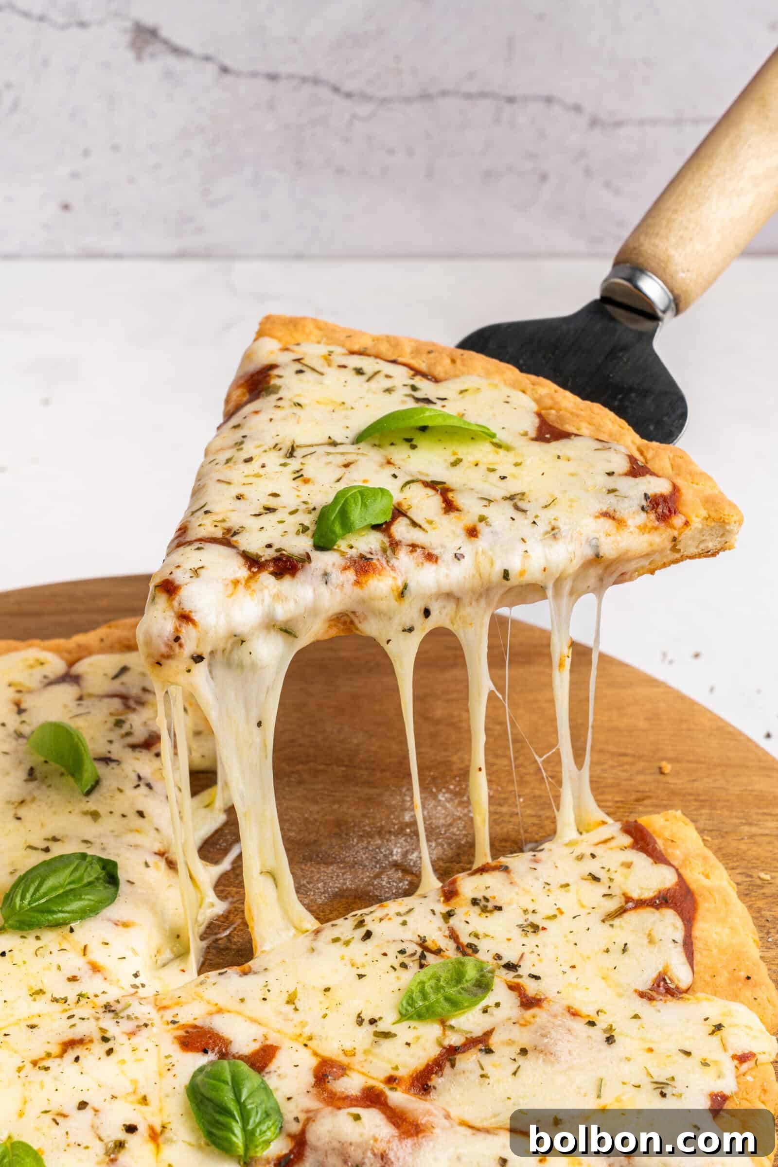 A hand lifts a cheesy slice of pizza from a wooden board, with melted cheese stretching between the slice and the pizza. The pizza is topped with fresh basil leaves and herbs.