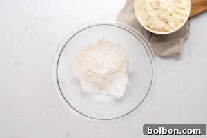 A glass mixing bowl with flour and baking powder sits on a white surface. Next to it is a bowl of ricotta cheese placed on a beige cloth on a wooden board.