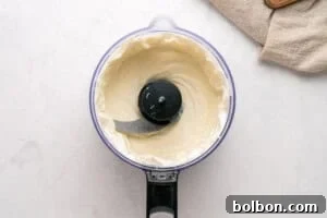 A food processor filled with smooth, creamy white mixture sits on a light countertop, with a beige kitchen towel partially visible in the upper right corner.
