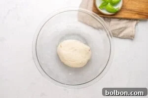 A ball of dough sits in a clear glass bowl on a white countertop. Nearby, a small plate with fresh basil leaves rests on a wooden board and beige cloth.