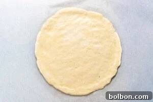 A round, uncooked pizza dough is rolled out flat on a sheet of parchment paper, ready for toppings and baking.