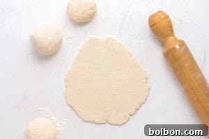 High-Protein Cottage Cheese Flatbreads 10 A rolled-out piece of dough is on a floured surface next to a wooden rolling pin and three round dough balls, ready for baking or cooking.