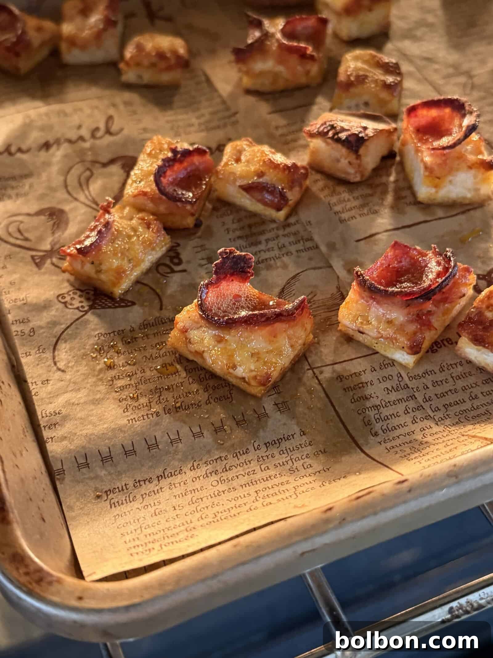 Small, square pieces of toasted bread topped with melted cheese and crispy bacon are arranged on a parchment-lined baking sheet inside an oven—perfect for adding crunch to a Pizza Crouton Salad. The parchment paper is printed with French text and illustrations.