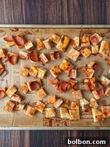 A baking sheet covered with parchment paper holds many small, square pizza croutons with various toppings, perfect for making a flavorful Pizza Crouton Salad, all spread out over a wooden surface.