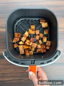 A hand holds an air fryer basket containing crispy, golden-brown cubes of tofu, perfect as a protein-packed topping for a Pizza Crouton Salad, displayed on a wooden surface.